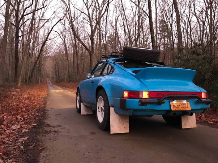 rear 3/4 view of a Custom Built 1980 Porsche 911 SC in Riviera Blue driving through the woods at dusk