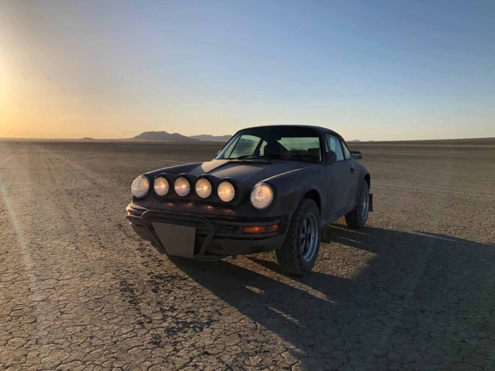 Custom Built 1987 Porsche 911 Carrera in Cassis Red with European bus fabric interior parked in front of a sunset with the headlights on