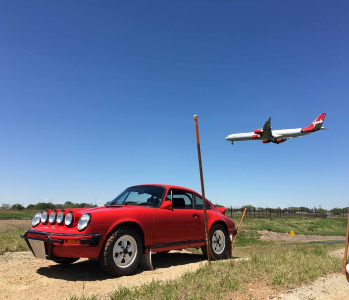 Custom Built 1981 Guards Red Porsche 911 SC parked in a field in front of an airplane