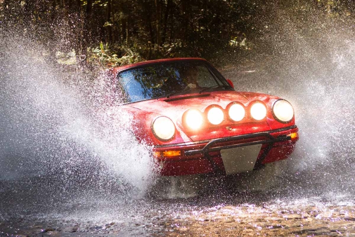 Custom Built 1981 Guards Red Porsche 911 SC with the headlights on driving through water